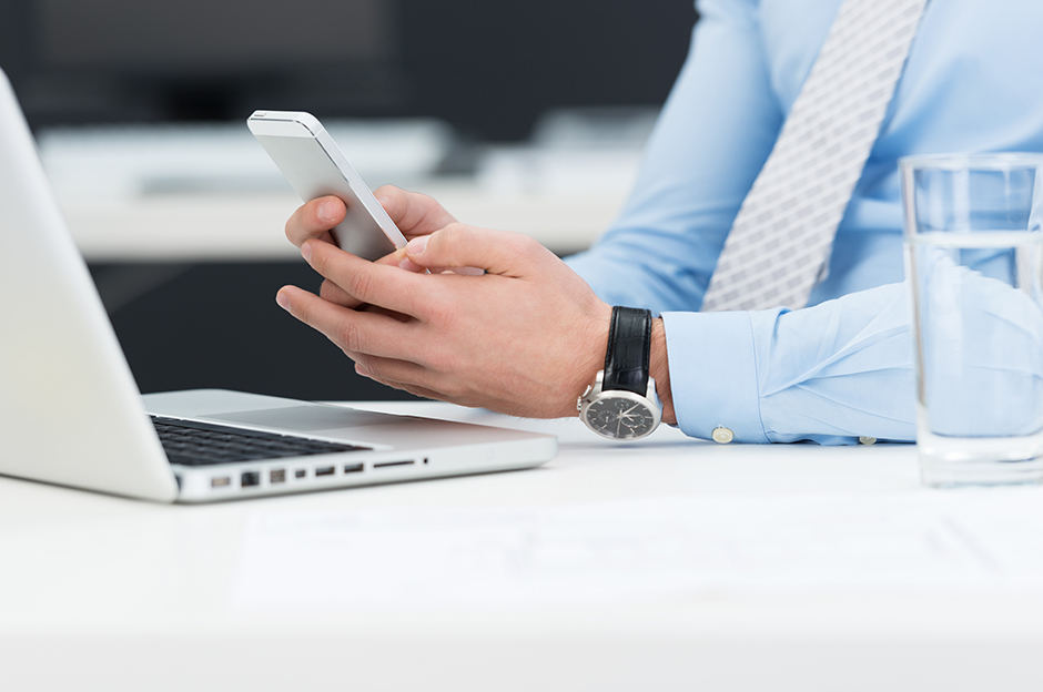 Businessman with tie holding a phone in one hand and sitting at a desk with a laptop on it.